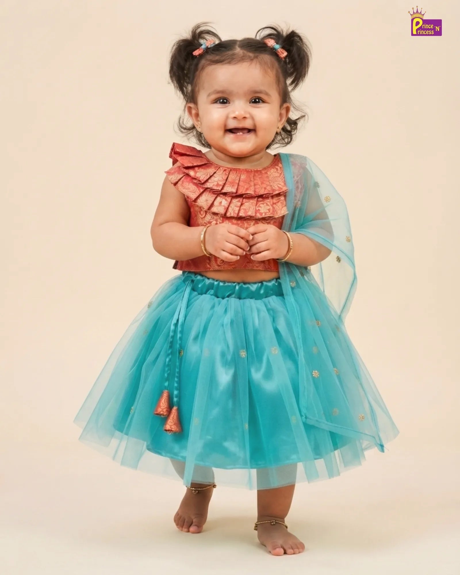 A toddler girl wearing a two-piece costume set consisting of a ruffled coral-orange top and a shiny turquoise-blue tutu skirt with gold polka dots and a matching sheer blue cape, standing against a solid beige background.