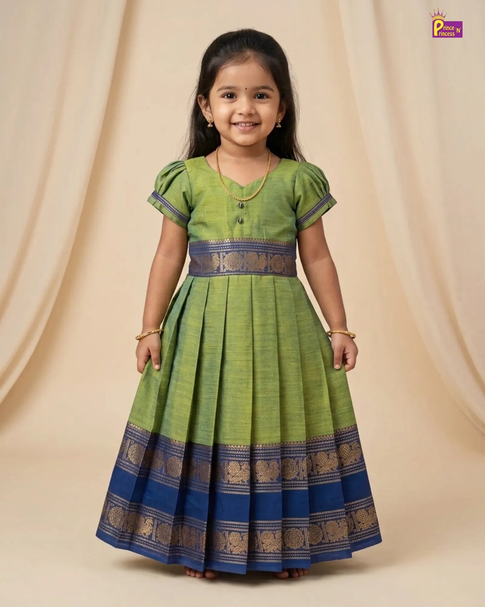 A young girl modeling a green and blue traditional Indian dress with gold zari borders, standing against a beige draped backdrop.