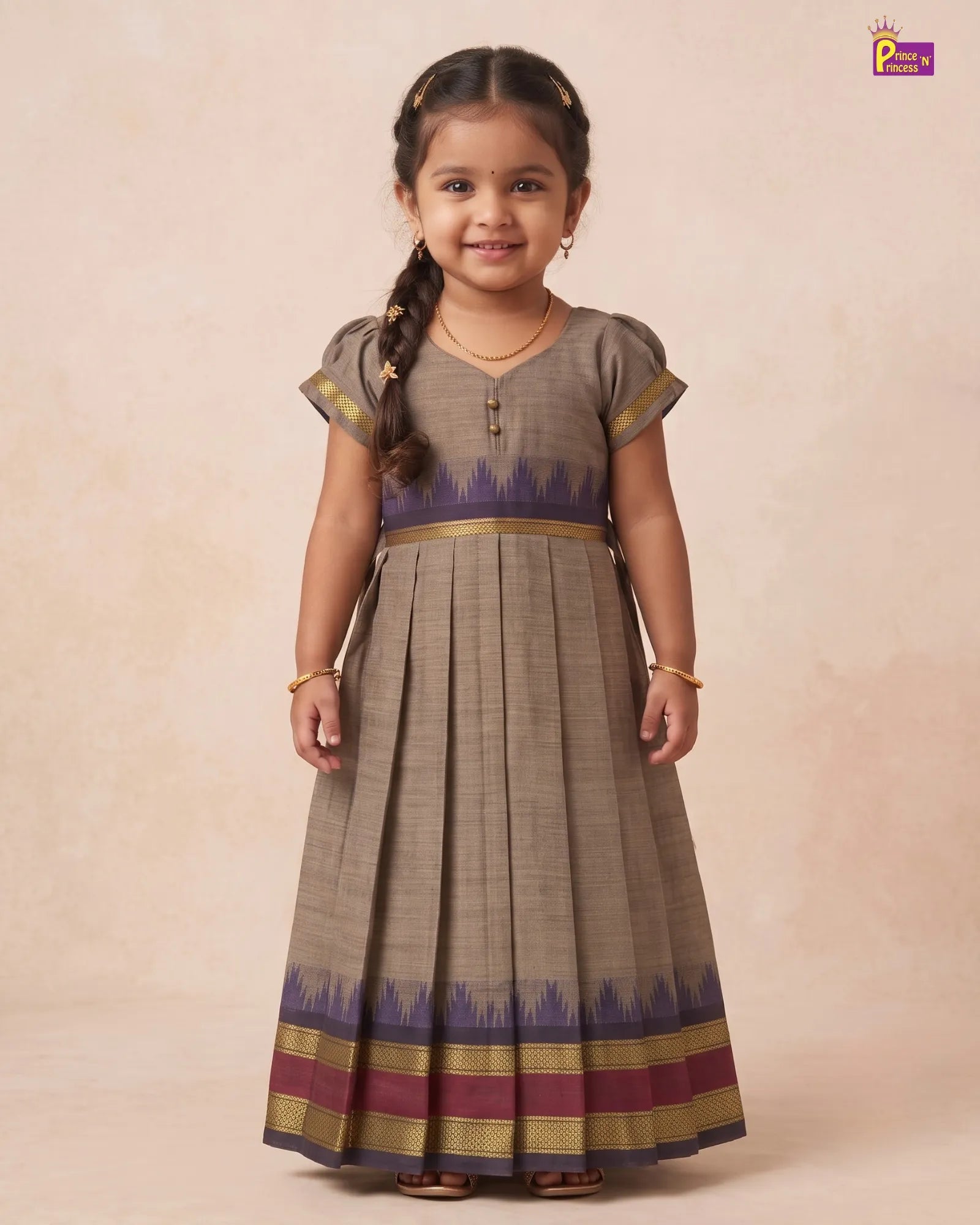 A young girl with a braid stands against a warm beige studio backdrop wearing a long pleated dress. The dress is taupe-brown with a purple and gold patterned hem and waistband.