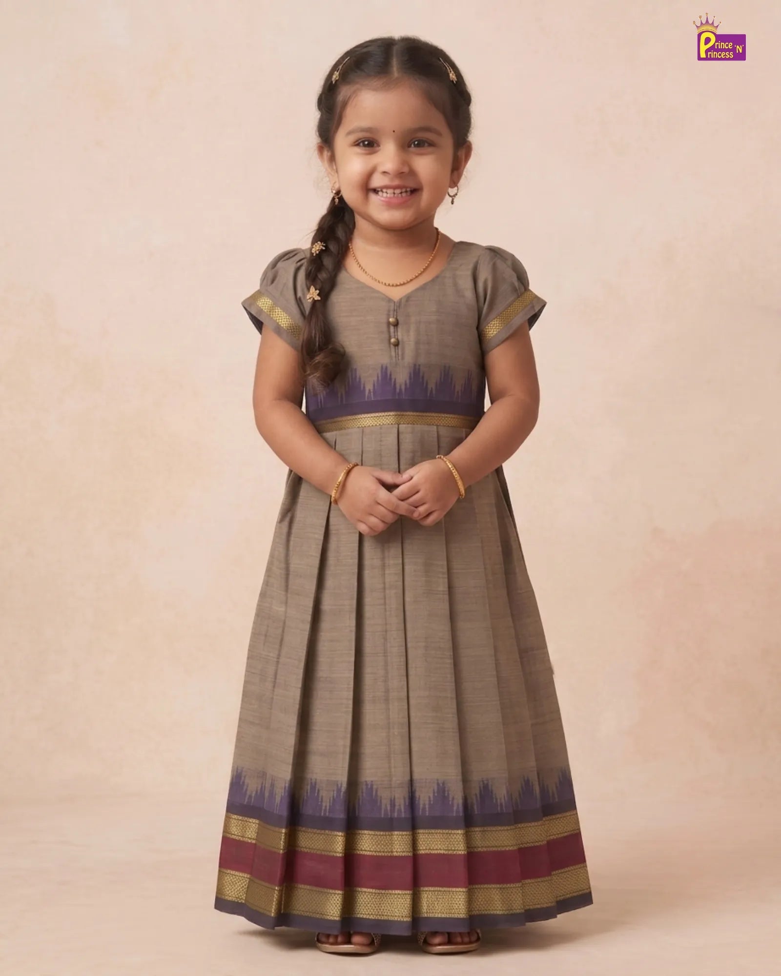 A young girl with braided hair stands wearing a traditional pleated Anarkali-style dress. The dress is primarily taupe brown with a purple and gold patterned waistband and hem, set against a warm beige studio backdrop.