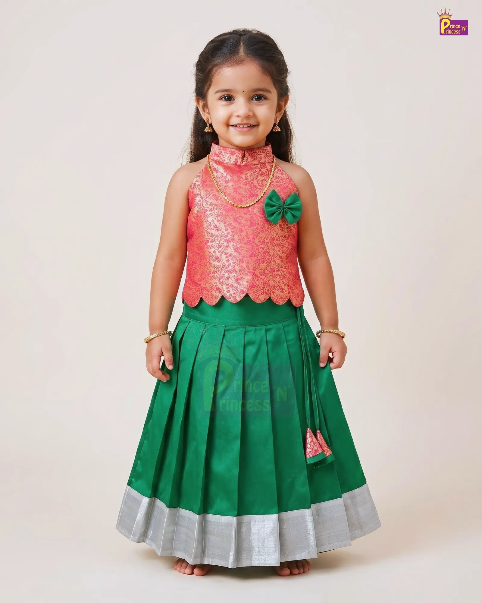 A young girl modeling a traditional Indian ethnic outfit consisting of a pink embroidered sleeveless top and a green pleated skirt with a silver border, set against a plain light beige background.