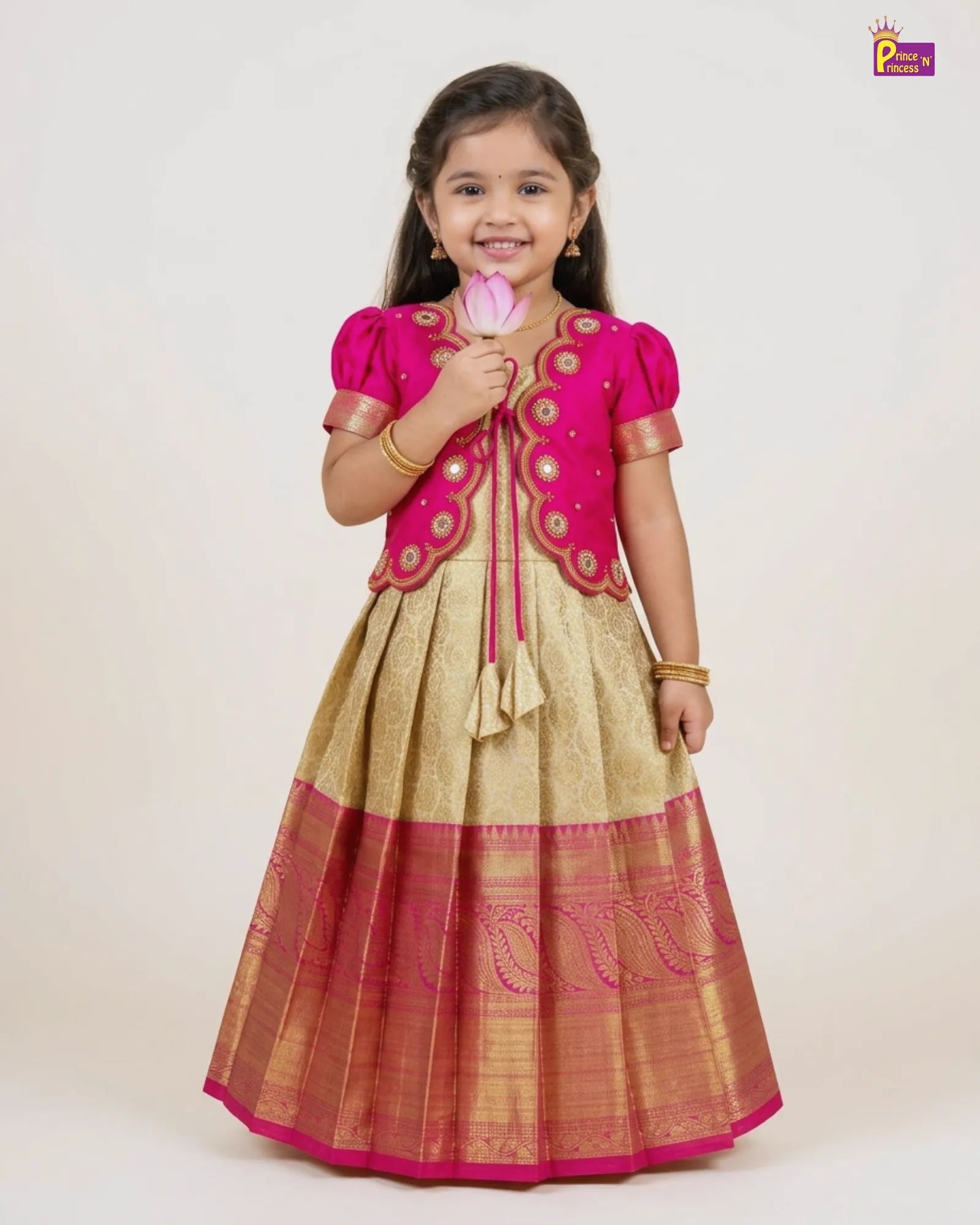 A young girl modeling a traditional Indian ethnic dress with a bright pink embroidered bodice and a gold-beige flared skirt with a pink border, standing against a plain light background.