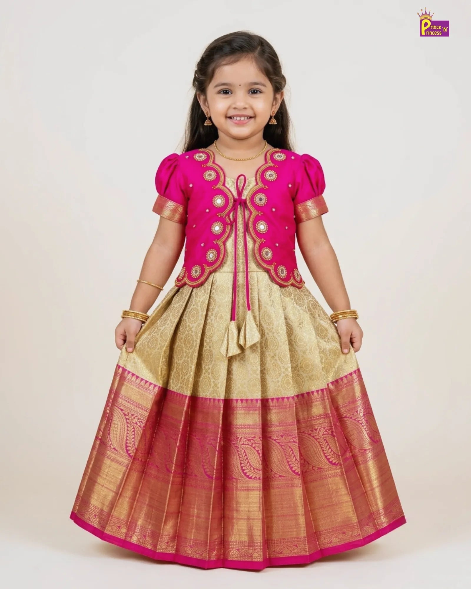 A young girl modeling a traditional Indian ethnic dress with a bright pink top and a gold and pink skirt, set against a plain light background.