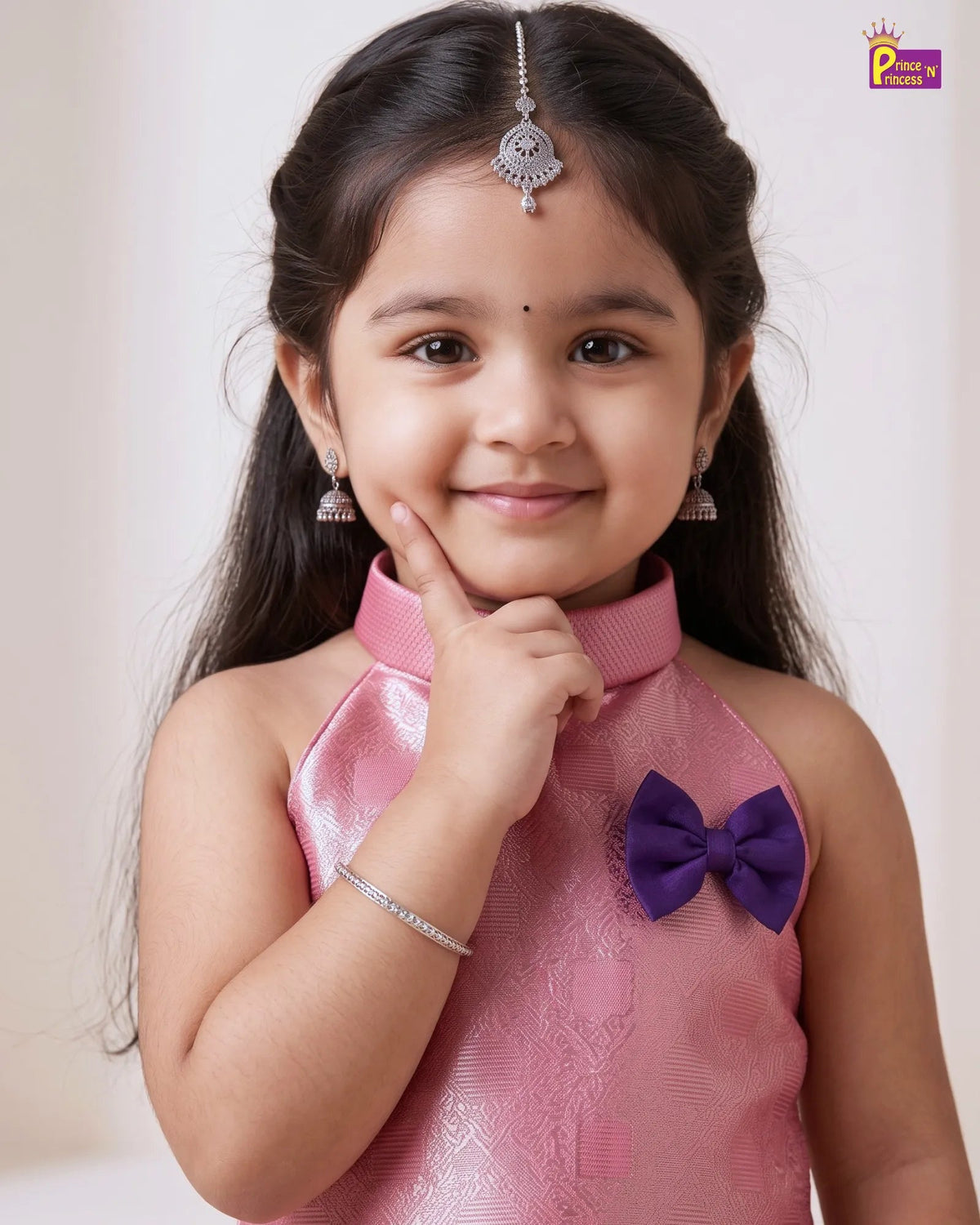 A young girl wearing a pink traditional Indian dress with a purple bow accent, silver earrings, and a silver maang tikka, posing against a light beige background.