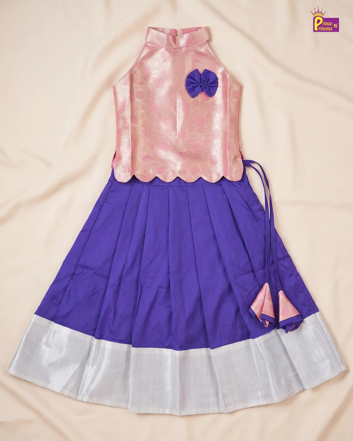 A girls' traditional Indian ethnic dress or lehenga choli set laid flat on a beige fabric background. The top is light pink with a scalloped hem and a purple bow accent, while the pleated skirt is purple with a white border and a matching pink tie detail.