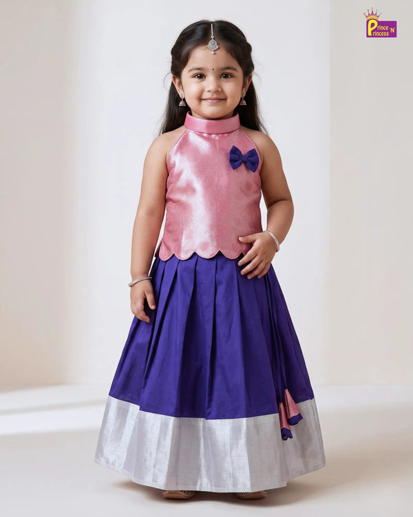 A young girl modeling a traditional Indian dress with a pink satin sleeveless top, a purple skirt, and a white border hem, standing against a plain light beige background.