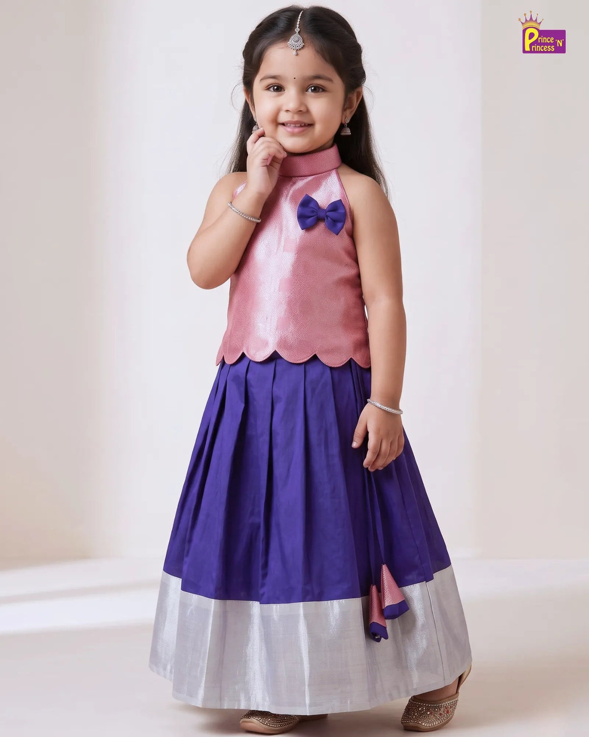 A young girl modeling a sleeveless traditional Indian dress with a pink satin bodice, purple bow accent, pleated purple skirt, and a wide silver lower border, set against a plain cream background.