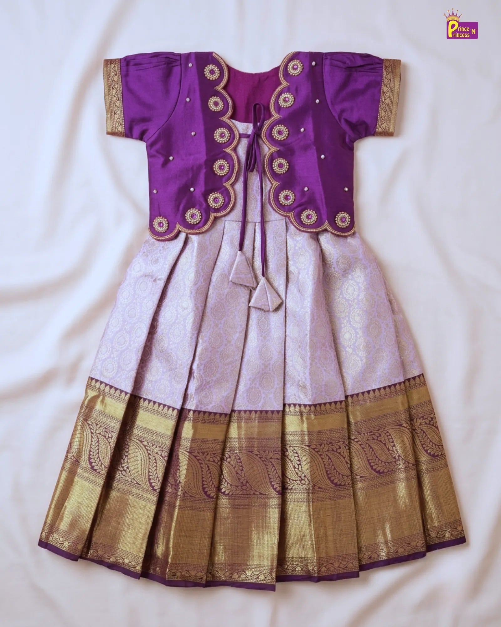 A traditional Indian girl's dress (lehenga choli) laid flat on a white fabric background. The choli is purple with gold and silver zari embroidery, and the skirt is light lavender with a wide gold zari pleated border.