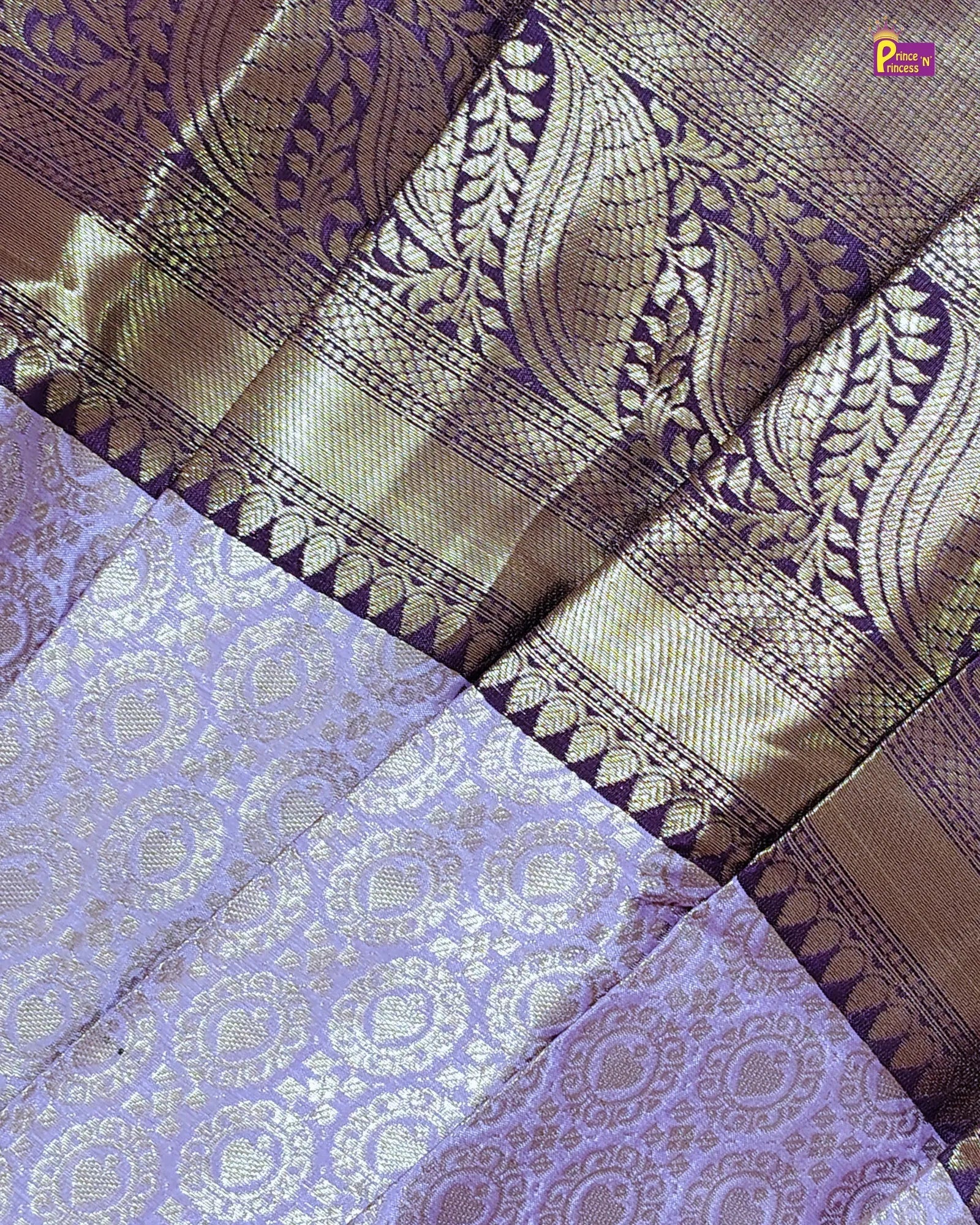 A close-up of a woven fabric, likely a sari, featuring a lavender purple body with metallic gold floral motifs and a wide gold zari border with matching paisley patterns.