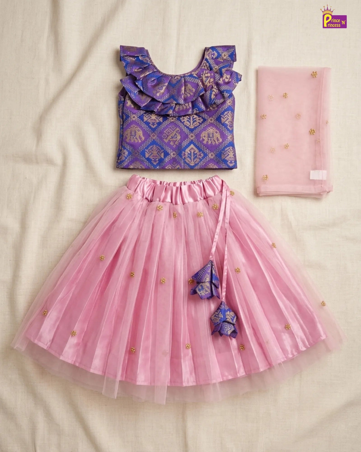 A girls' two-piece ethnic outfit set laid flat on a cream fabric background, featuring a purple patterned ruffled top and a pink tulle skirt with gold embroidered motifs and a matching pink dupatta.