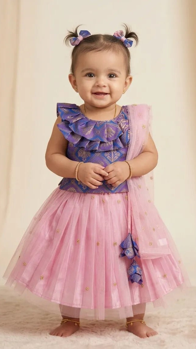A toddler girl wearing a two-piece dress with a purple patterned ruffled top and a pink tulle skirt with gold star details, standing against a beige draped backdrop.