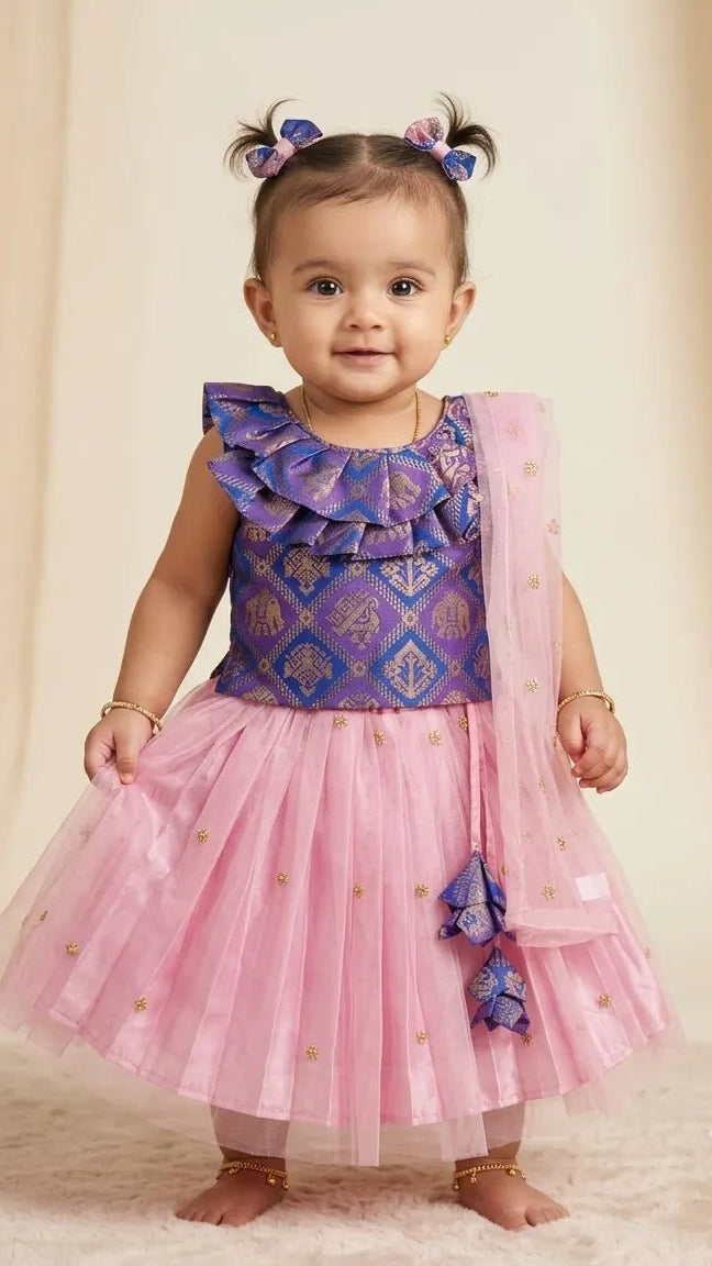 A toddler girl wearing a purple and pink ethnic party dress with a patterned bodice, ruffled shoulder details, a pink tulle skirt with gold foil dots, and a matching sheer pink dupatta, standing against a beige draped backdrop.