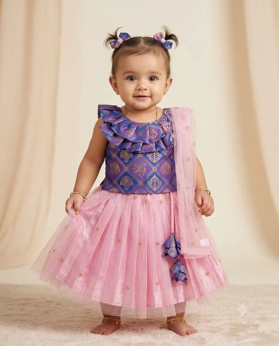 A toddler girl wearing a purple and pink ethnic party dress with a patterned bodice, ruffled shoulder details, a pink tulle skirt with gold foil dots, and a matching sheer pink dupatta, standing against a beige draped backdrop.