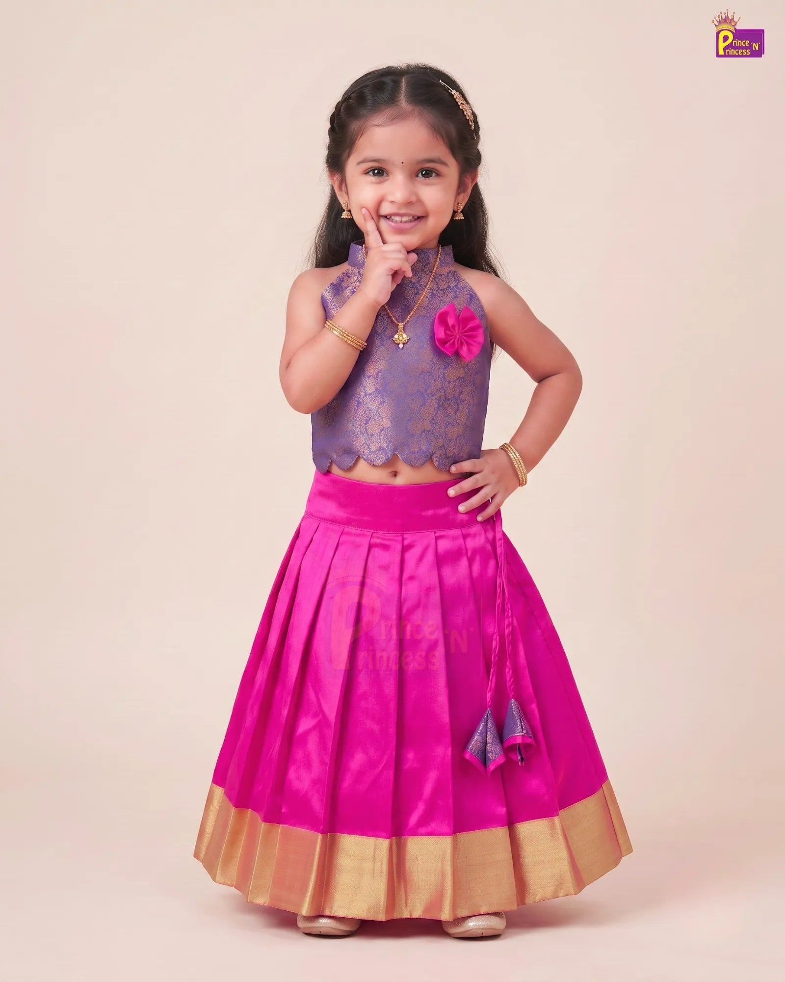 A young girl modeling a traditional Indian ethnic outfit consisting of a purple embroidered sleeveless top and a bright pink pleated skirt with gold zari borders, set against a solid light beige background.