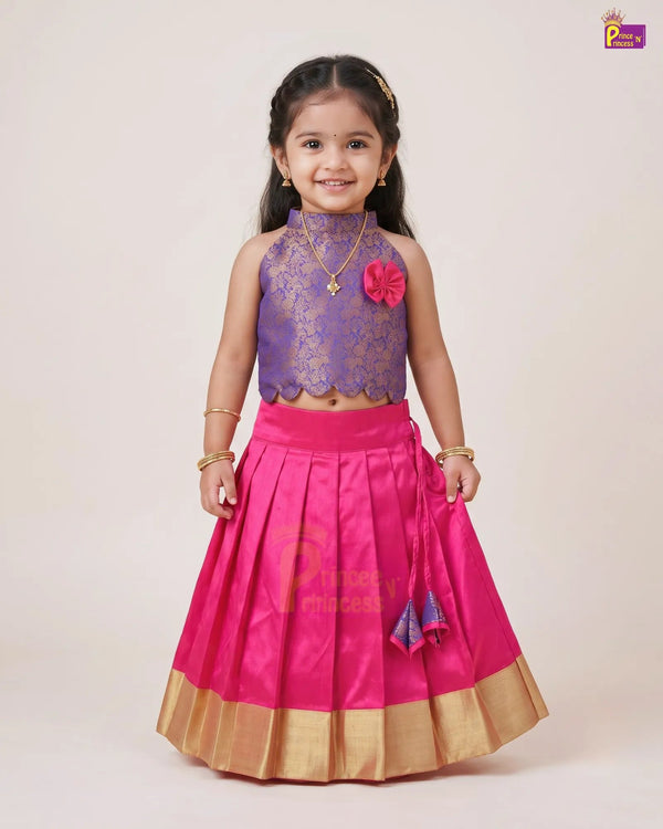 A young girl modeling a traditional Indian outfit consisting of a purple lace sleeveless crop top and a bright pink pleated skirt with gold borders, set against a plain beige studio background.