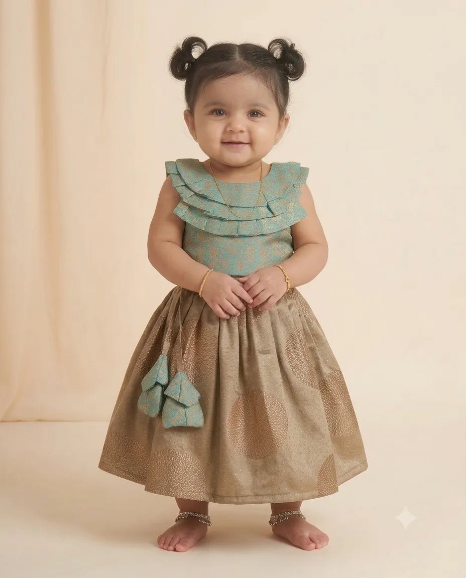 A toddler girl with two buns of hair stands barefoot against a soft beige studio backdrop, wearing a sleeveless traditional-style dress with a ruffled mint green bodice and a metallic gold skirt, plus a matching mint green bow detail on the skirt.