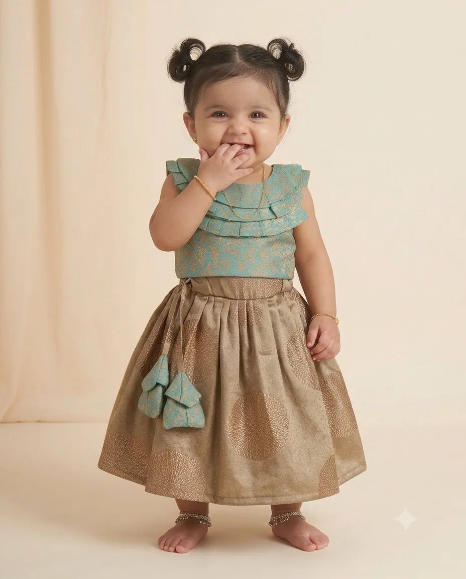 A toddler girl wearing a traditional Indian outfit consisting of a light teal ruffled sleeveless top and a metallic gold skirt with a matching teal bow, standing against a warm beige background.