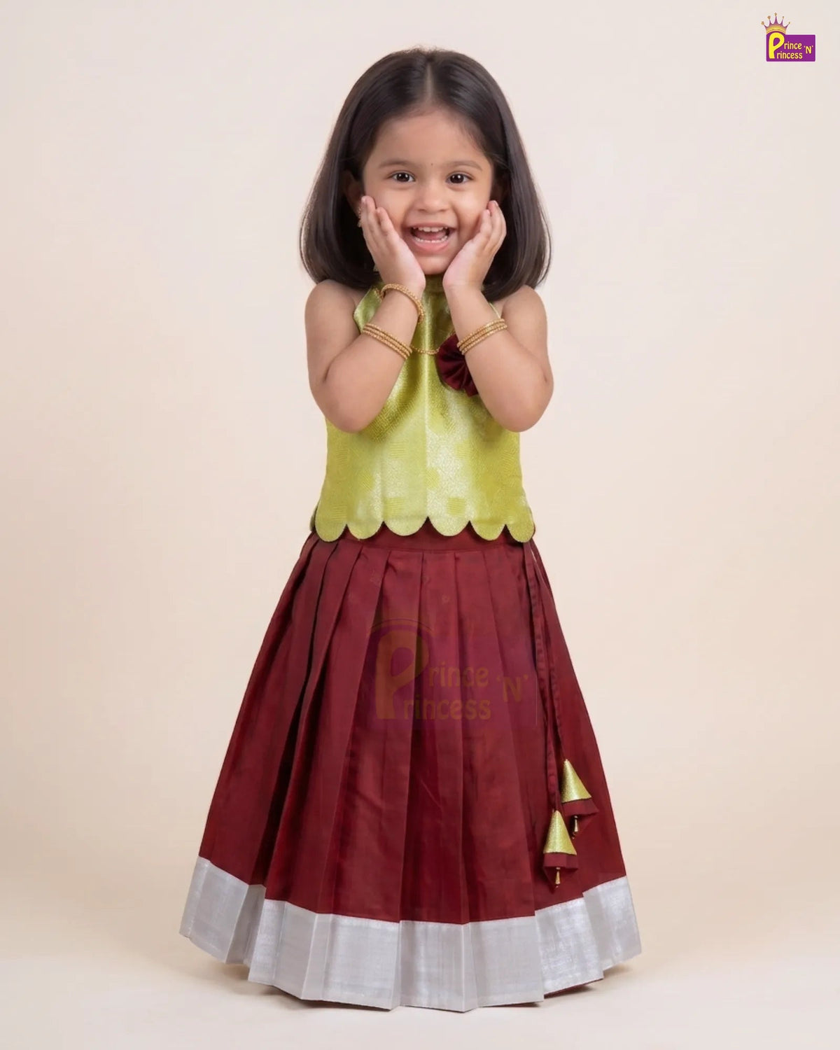 A young girl modeling a traditional Indian outfit consisting of a maroon skirt with a silver border and gold tassel detail, paired with a lime green sleeveless top, against a solid beige background.