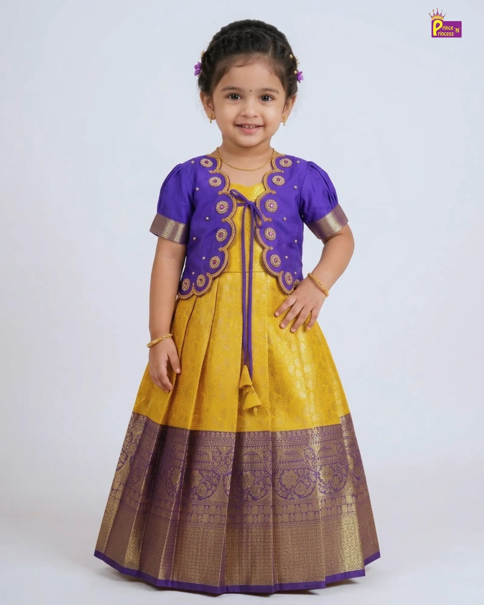 A young girl modeling a traditional Indian ethnic dress consisting of a yellow skirt with a wide purple and gold border, a matching yellow top, and a purple embroidered vest.