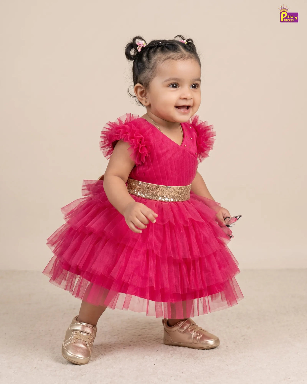 A toddler girl wearing a bright pink tiered tulle dress with a gold sequined waistband and metallic pink shoes, standing against a plain beige studio background.