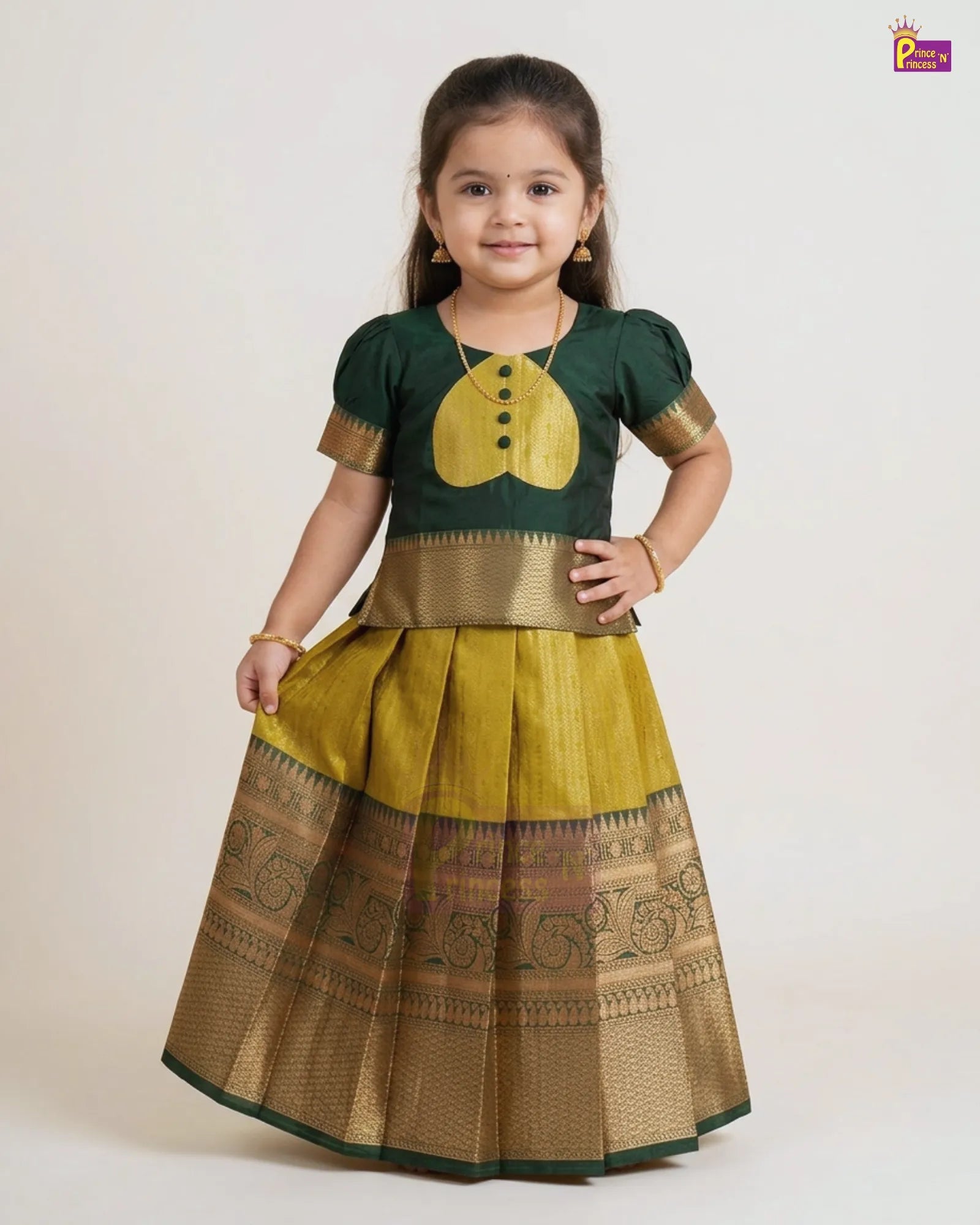 A young girl modeling a traditional Indian ethnic wear set consisting of a dark green short-sleeve blouse with a yellow chest motif and gold trim, paired with a pleated yellow skirt featuring a wide gold zari border.