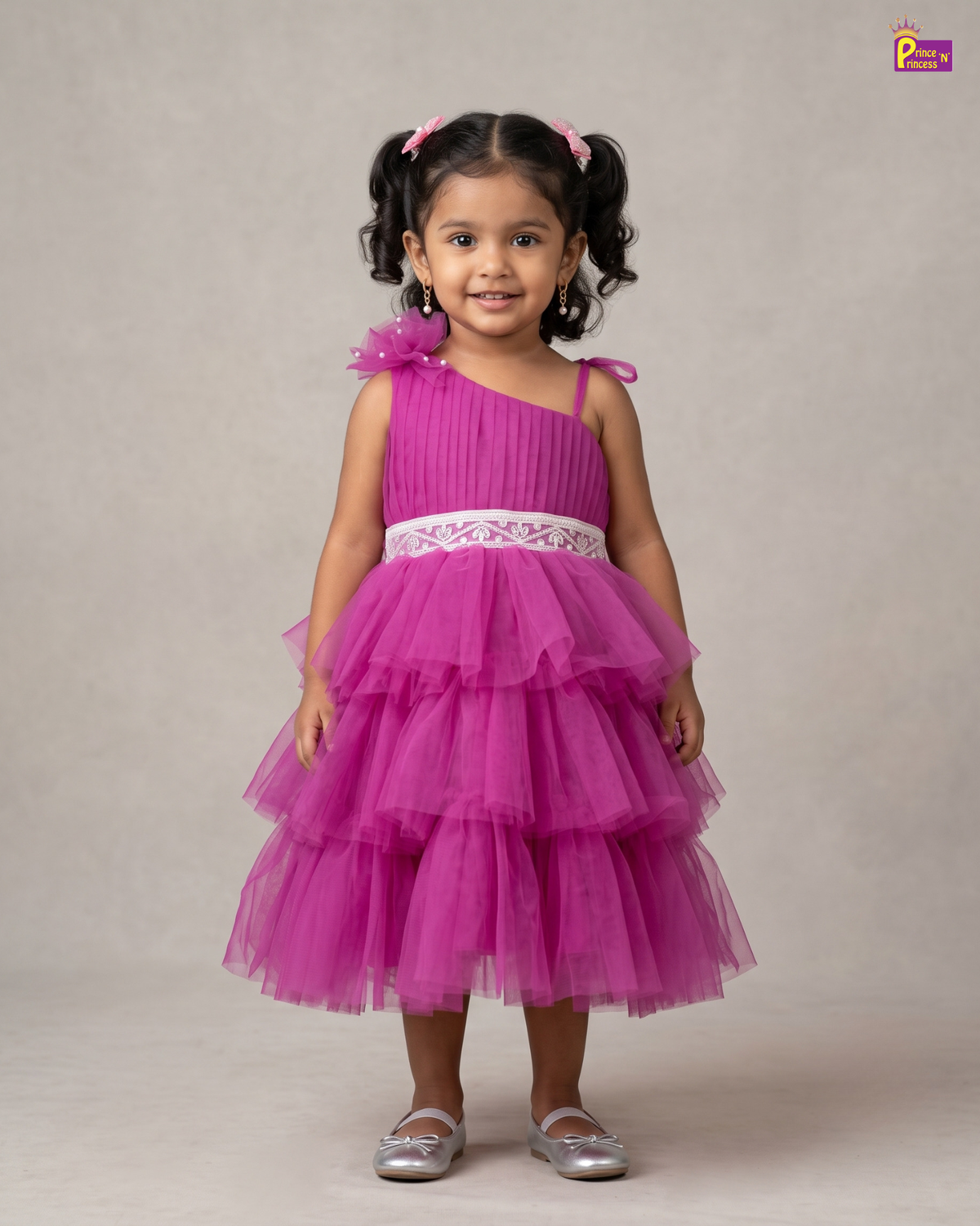 A young girl with dark curly hair in pigtails wearing a bright magenta pink one-shoulder tulle dress with a white lace waistband, standing against a neutral beige studio backdrop.