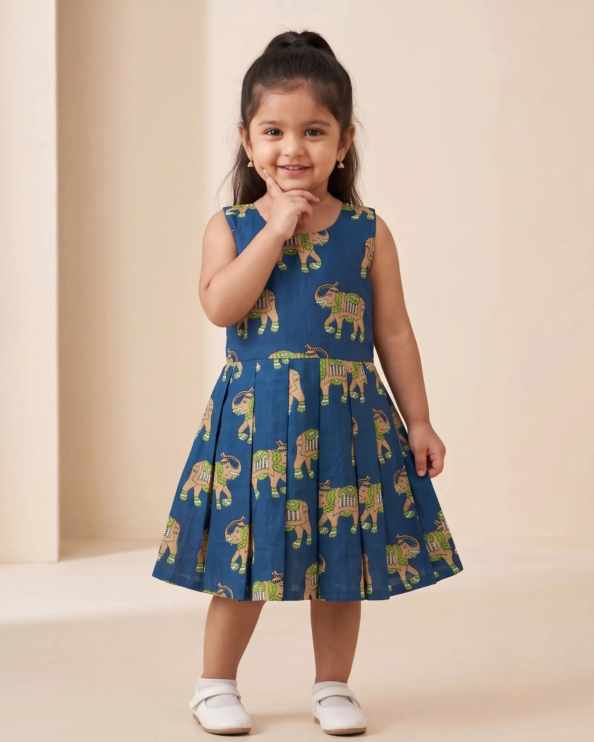 A young girl with her hair in a high ponytail stands wearing a sleeveless blue dress with a pattern of elephants and white shoes, posing in a studio with a beige background.