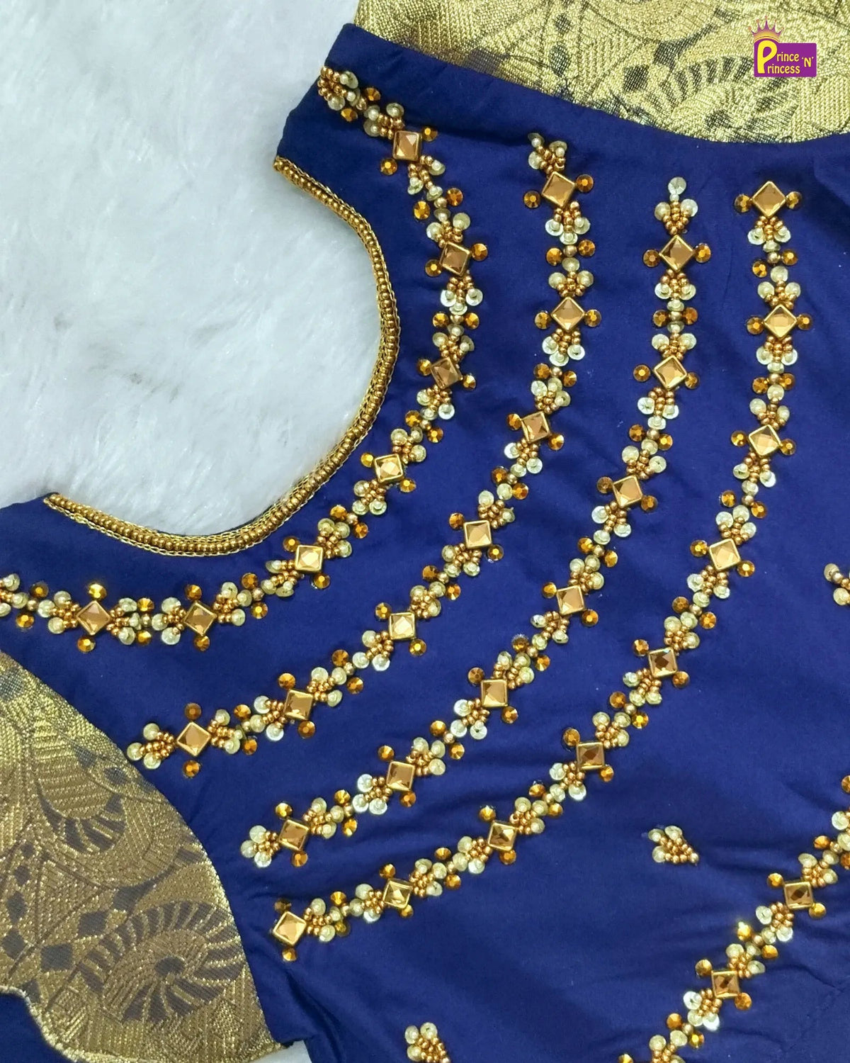 A close-up of a royal blue fabric garment with ornate gold and white beaded embroidery and a gold patterned border, laid flat on a white textured surface.