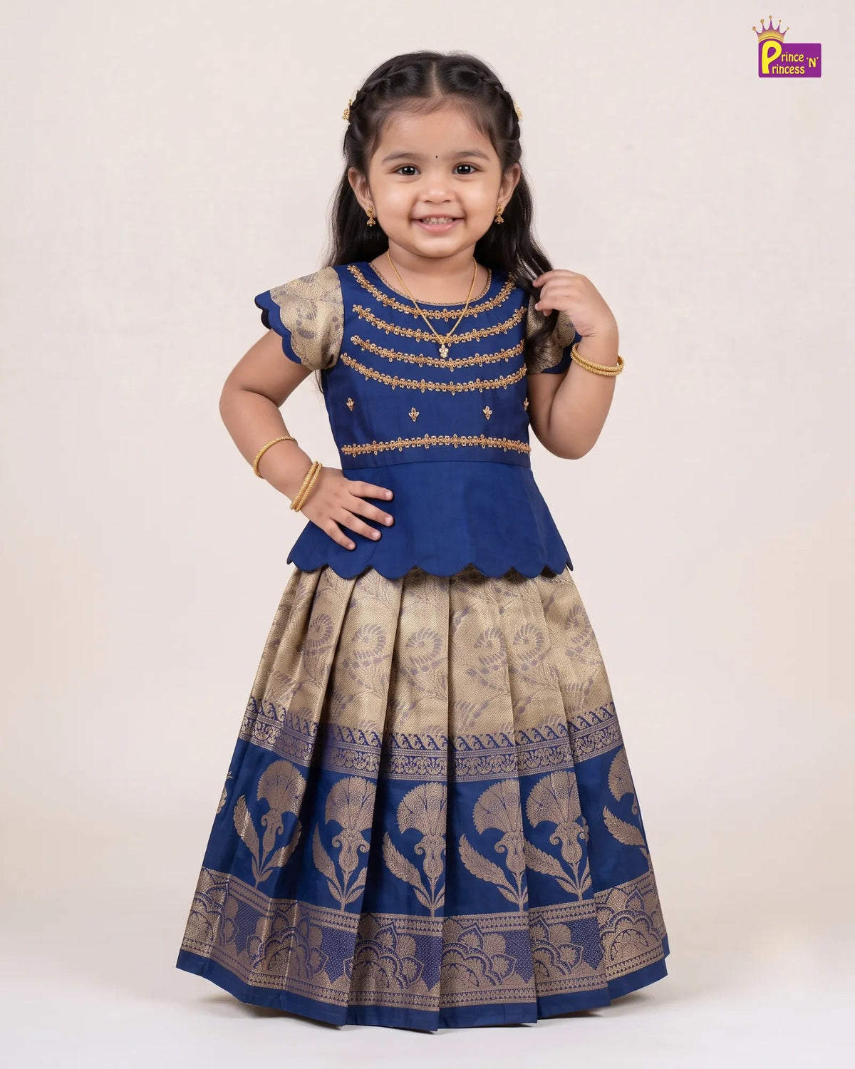 A young girl modeling a royal blue and beige printed traditional Indian dress with gold embroidery on the bodice, set against a plain cream background.