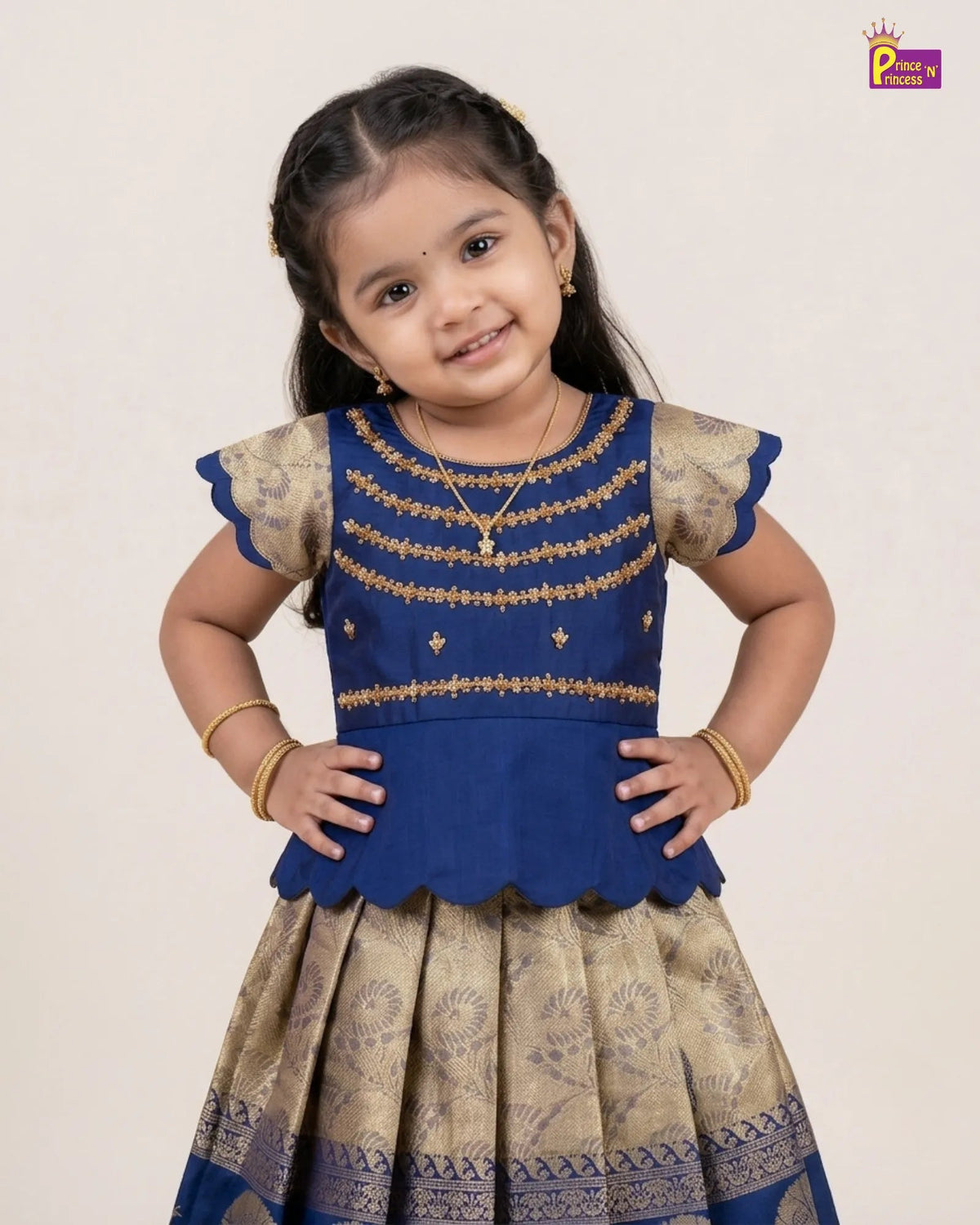 A young girl modeling a royal blue and beige printed traditional Indian dress with gold embroidery, standing against a plain light beige background.