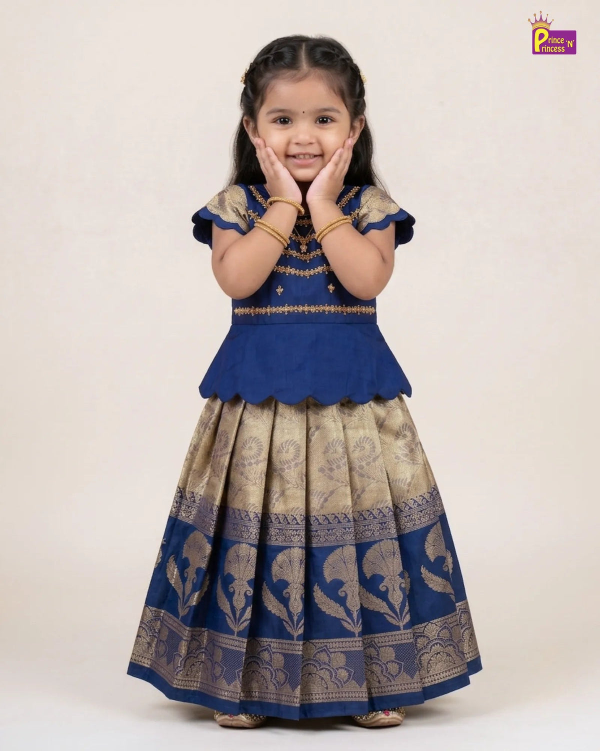 A young girl modeling a traditional Indian ethnic outfit consisting of a royal blue peplum top and a matching patterned flared skirt in beige and blue, set against a plain beige studio background.