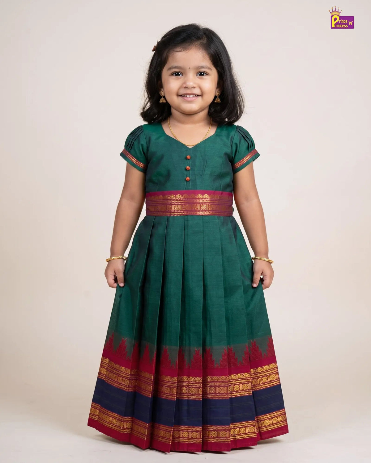 A young girl modeling a dark green traditional Indian dress with a red and gold border, standing against a plain beige studio background.