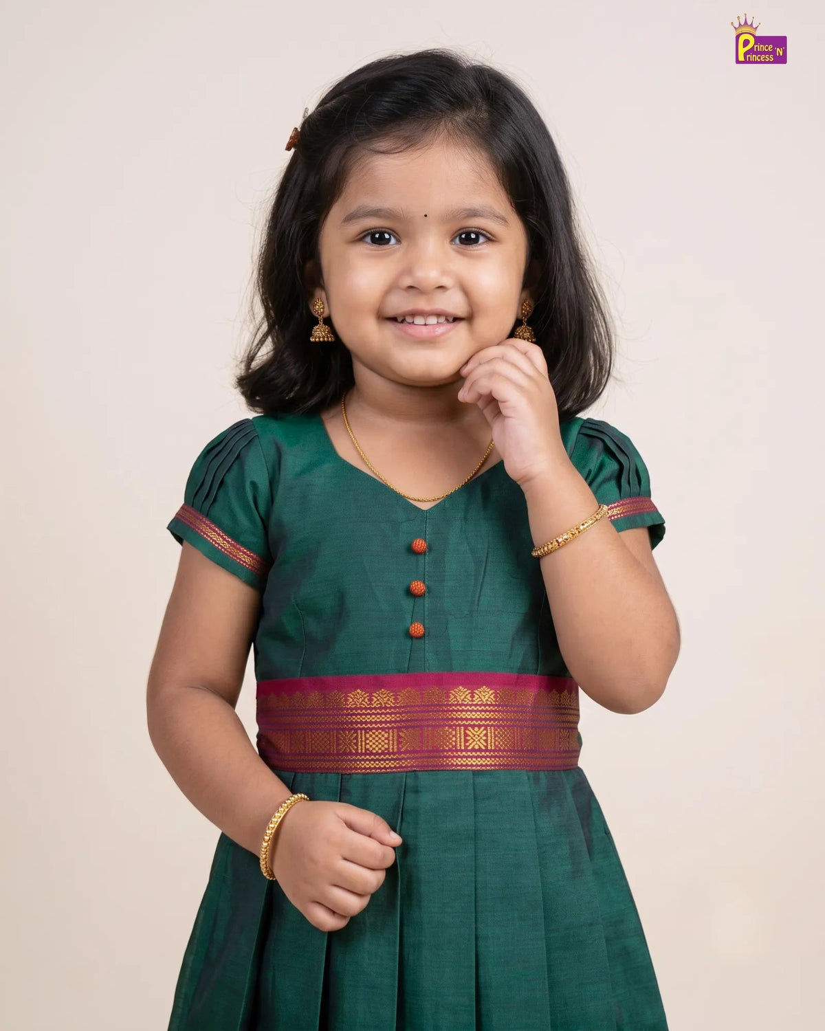 A young girl with dark hair is smiling and posing in a dark green traditional dress with a magenta and gold waistband, set against a plain beige background.