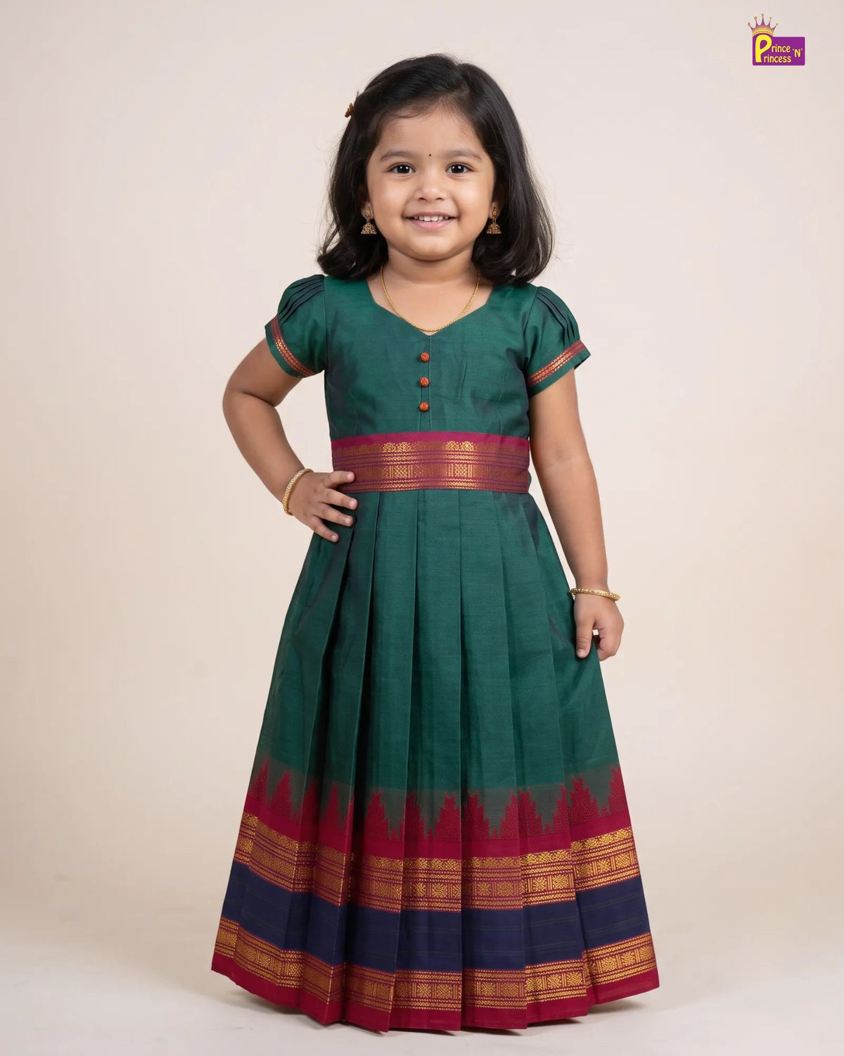 A young girl modeling a dark green traditional dress with a red and gold border, standing against a plain beige background.