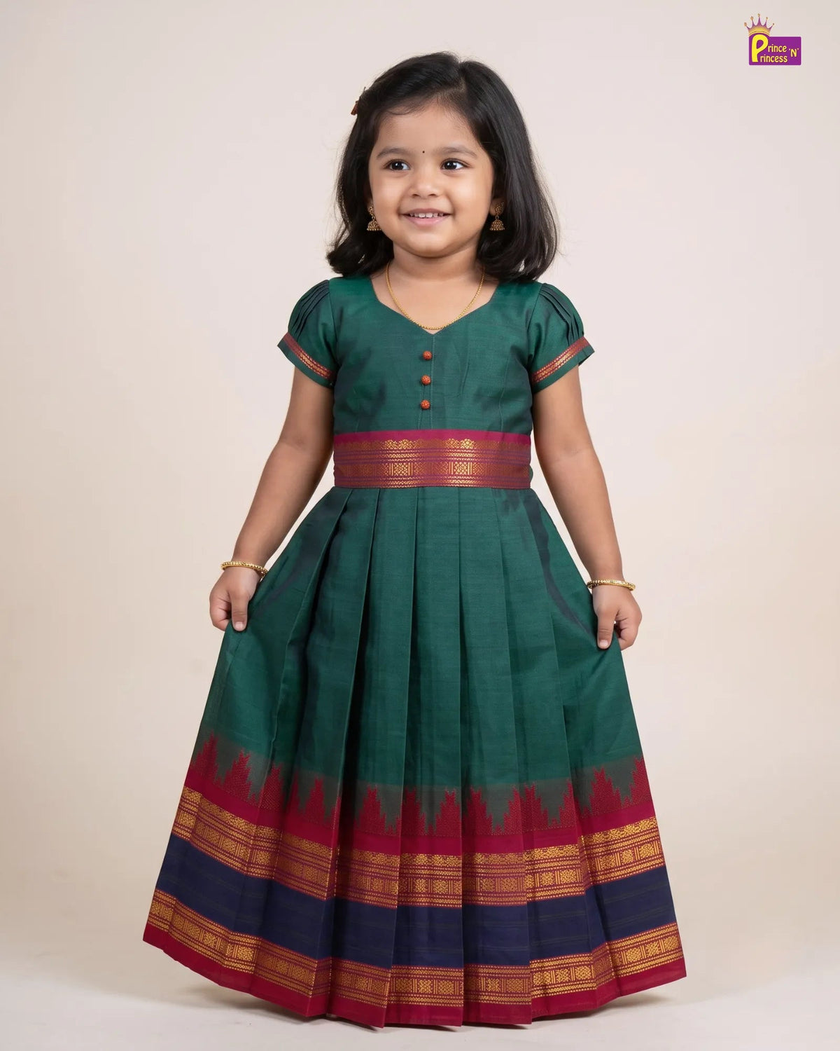 A young girl modeling a dark green traditional Indian dress with a red and gold border, standing against a plain beige background.