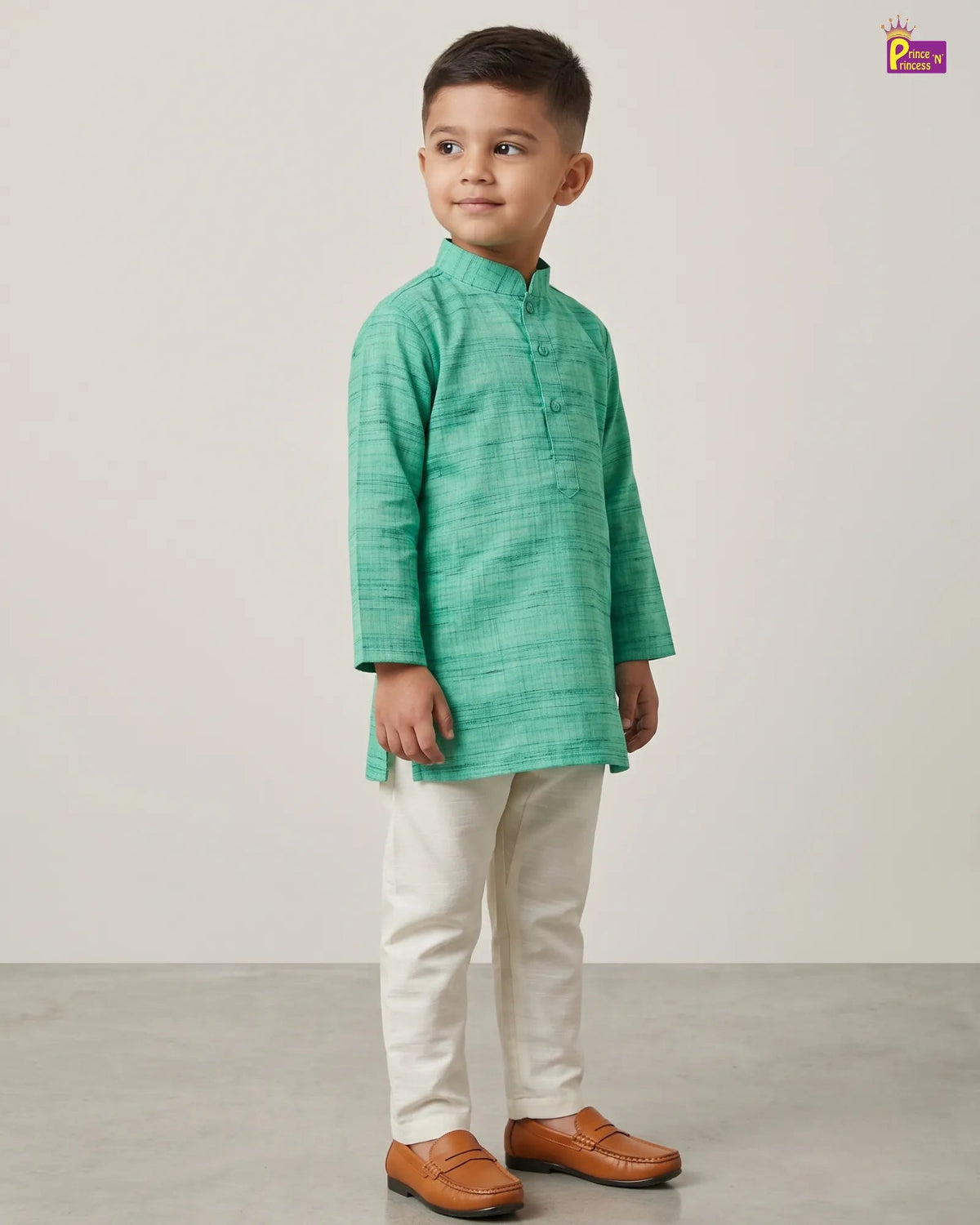 A young boy modeling a green textured kurta with cream trousers and brown leather shoes, standing against a neutral studio backdrop.