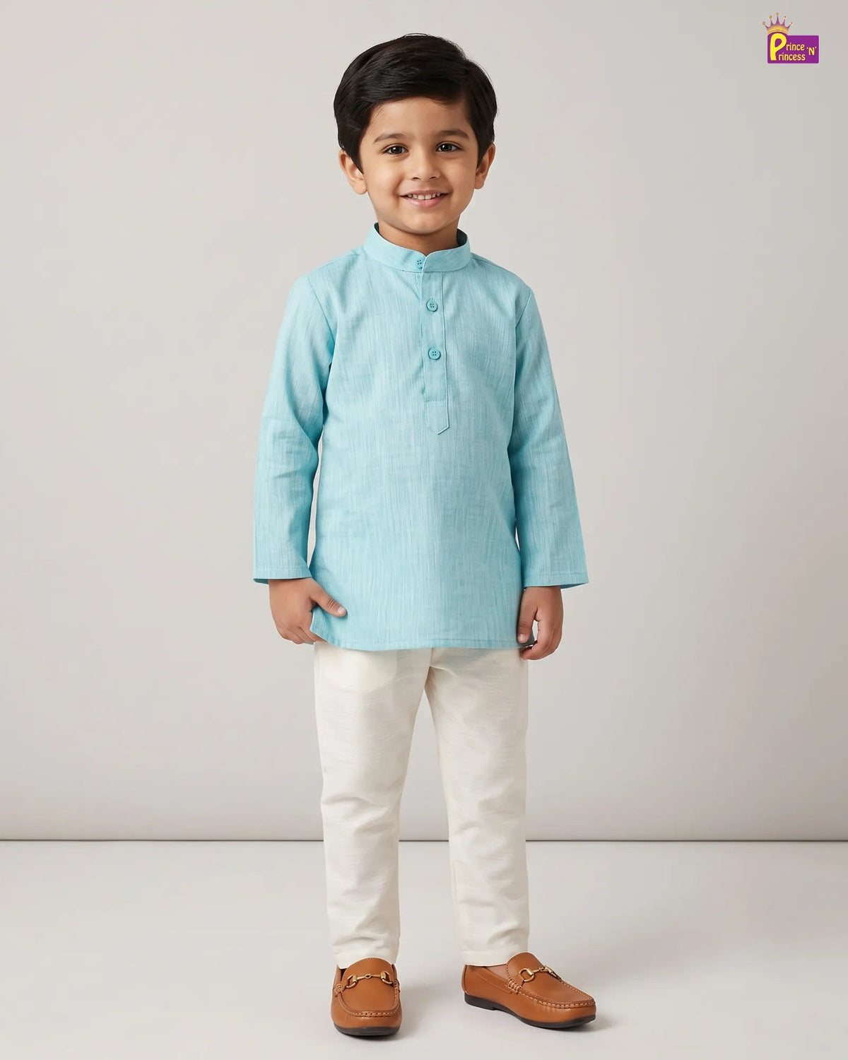 A young boy modeling a light blue long-sleeve kurta with cream trousers and brown loafers, standing against a plain light gray studio background.