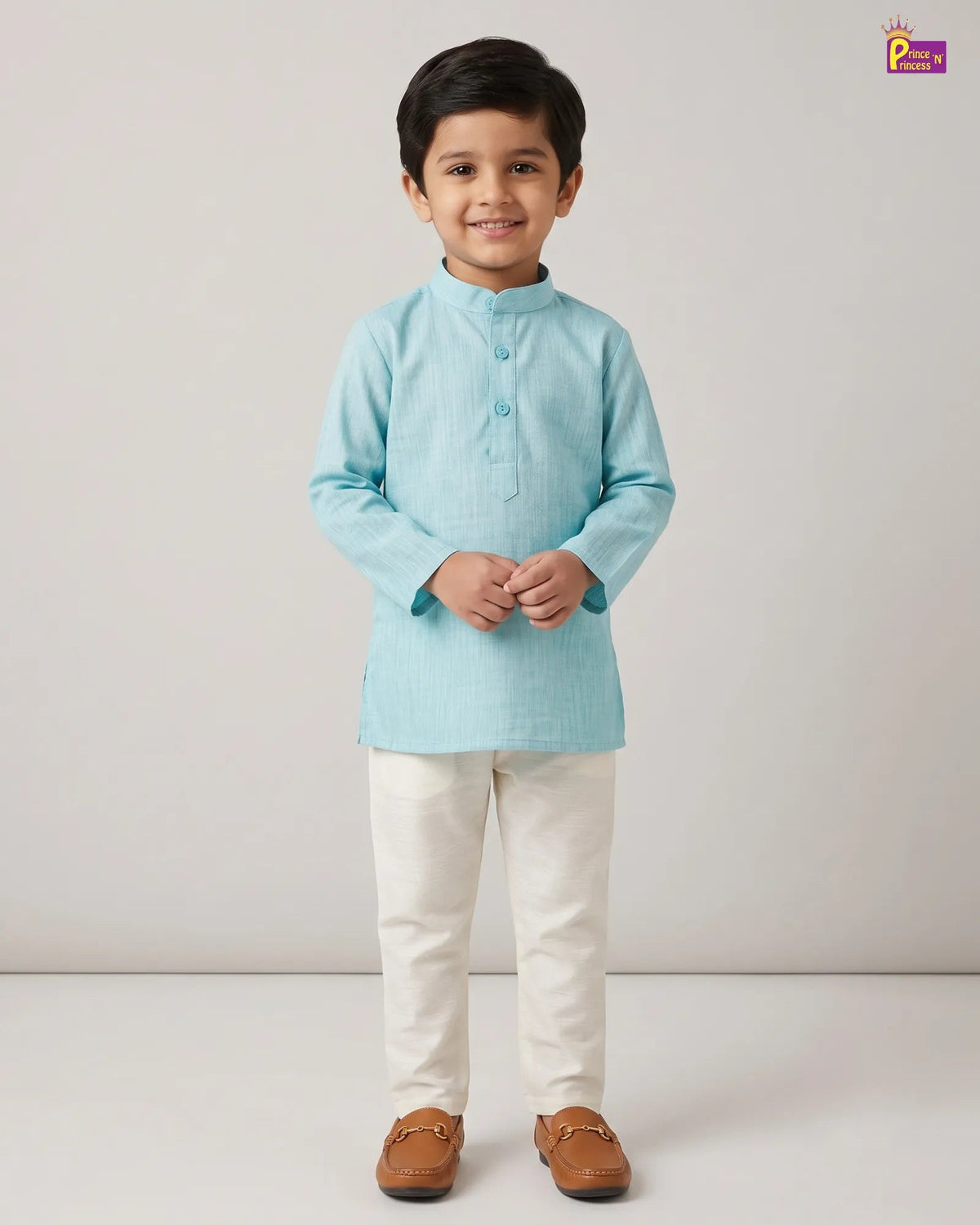 A young boy modeling a light blue long-sleeve kurta with cream trousers and brown loafers, standing against a plain light gray studio background.