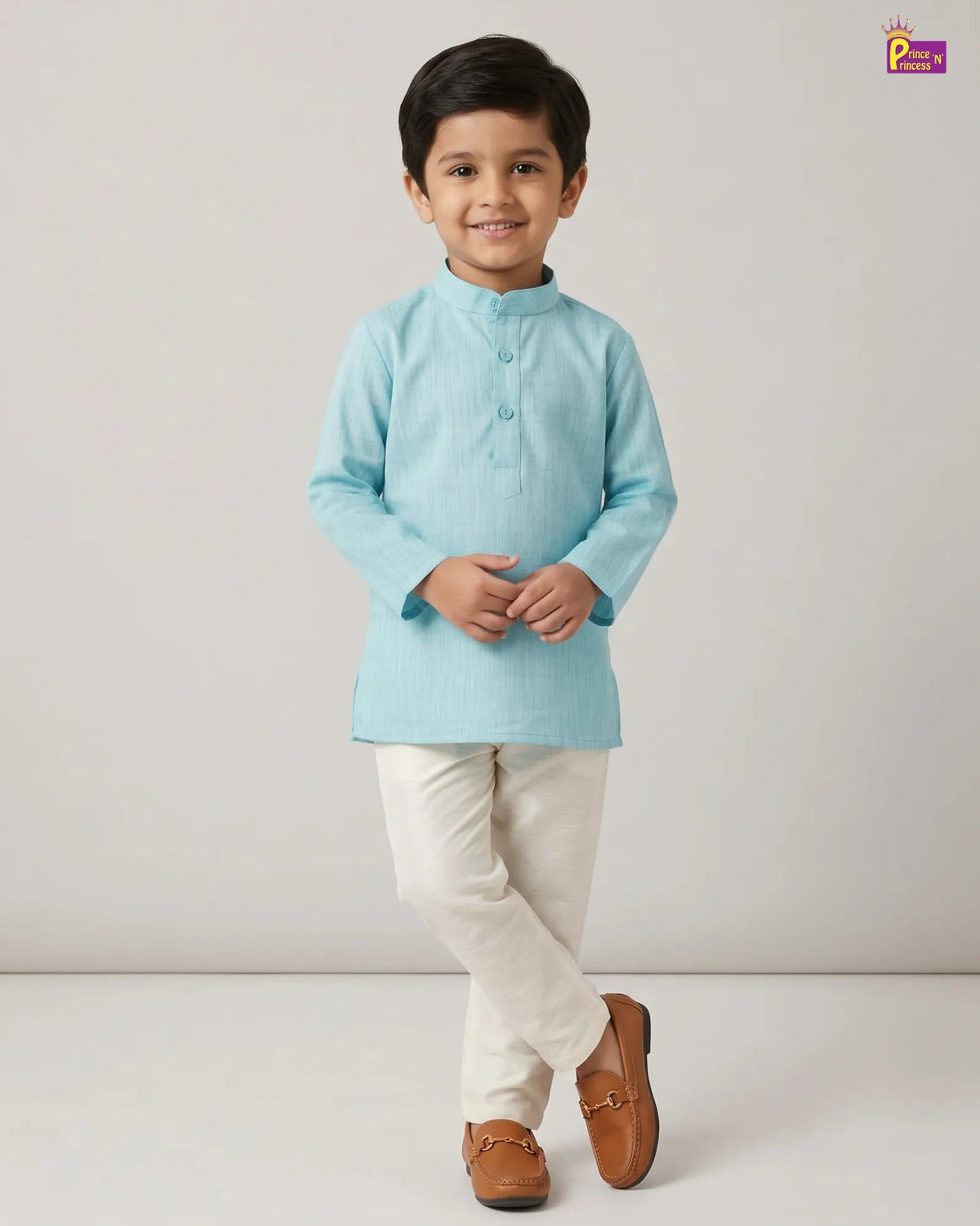 A young boy modeling a light blue long-sleeve kurta with cream trousers and brown loafers, standing against a neutral light gray studio background.
