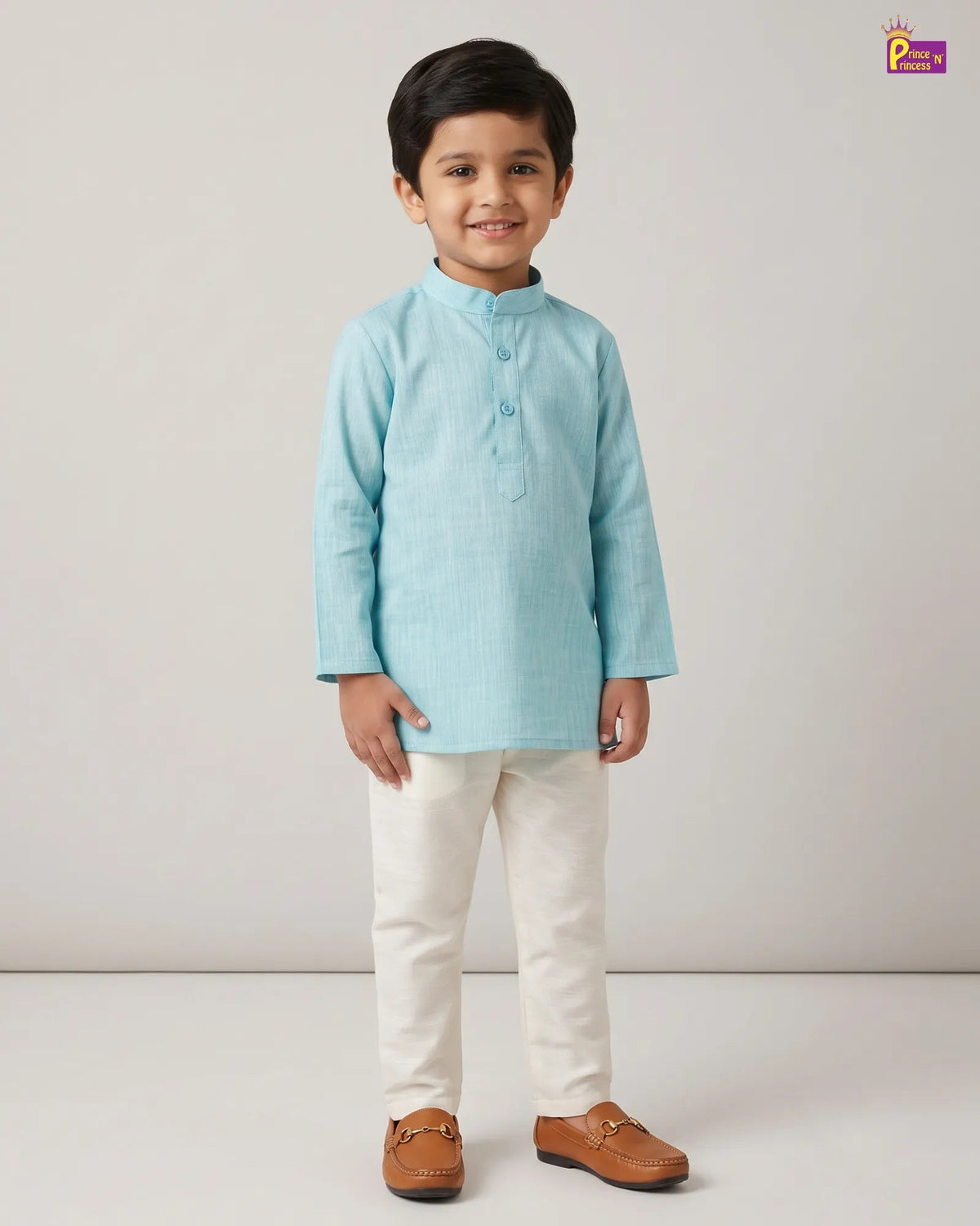 A young boy modeling a light blue long-sleeve kurta with cream trousers and brown leather loafers, standing against a neutral light gray studio background.