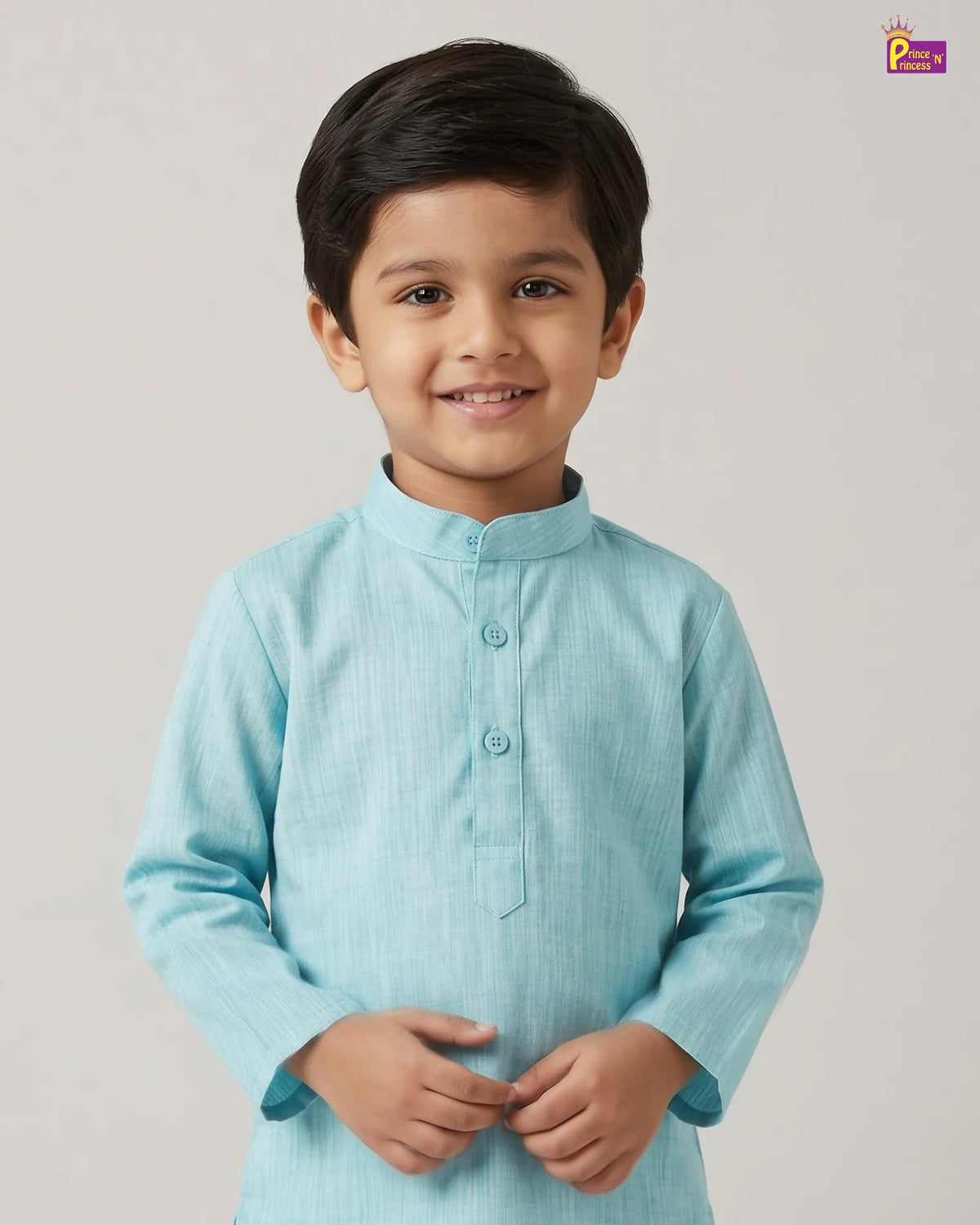 A young boy smiling and wearing a light blue long-sleeve kurta with a mandarin collar and three front buttons, photographed against a plain light gray studio background.