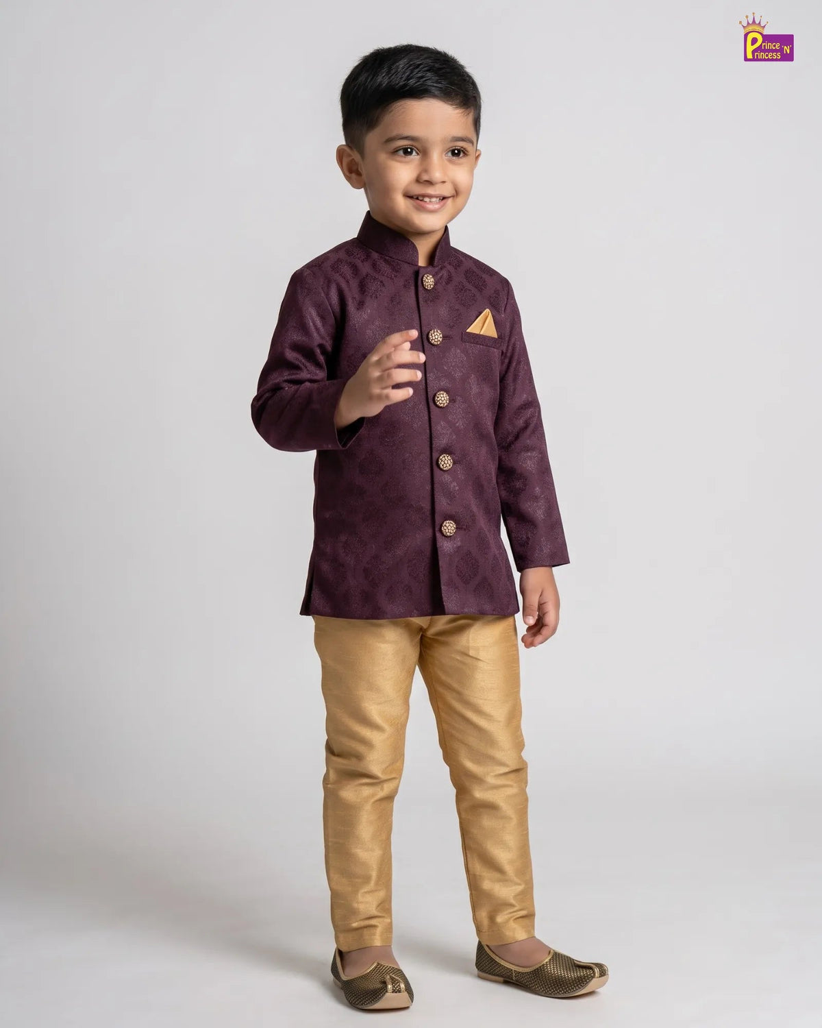 A young boy modeling a traditional Indian outfit consisting of a deep purple embroidered jacket with gold buttons and a matching gold pocket square, paired with gold trousers and brown shoes, against a light gray background.