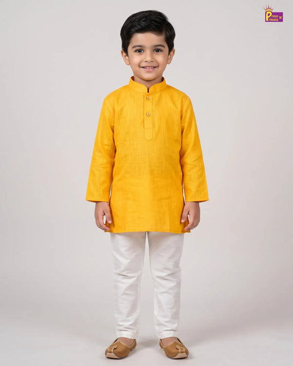 A young boy modeling a bright yellow long-sleeve kurta with white trousers and brown leather sandals, standing against a neutral light gray studio background.
