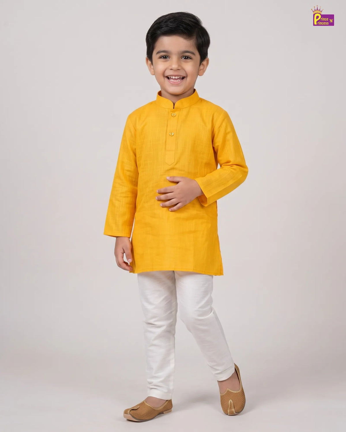 A young boy modeling a bright yellow long-sleeve kurta with white trousers and brown shoes, standing against a plain light gray studio background.