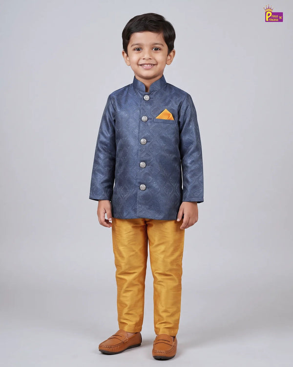 A young boy modeling a traditional Indian outfit consisting of a textured blue waistcoat jacket with a yellow pocket square, mustard yellow trousers, and brown leather shoes, shot against a neutral gray background.