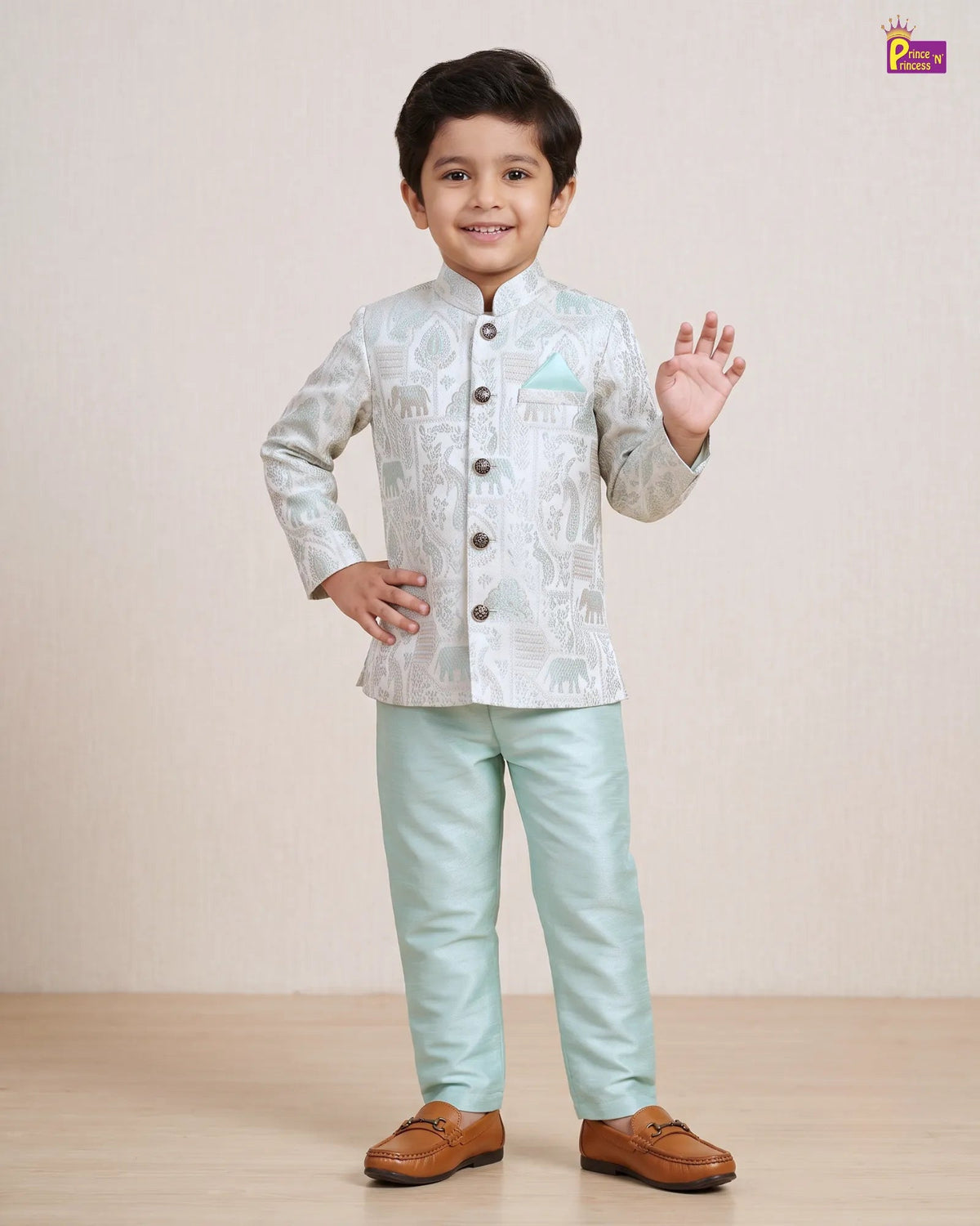 A young boy modeling a traditional Indian outfit consisting of a light sage green printed sherwani jacket with brown buttons and a matching light green trouser, paired with brown leather loafers. He is standing against a neutral beige studio backdrop.