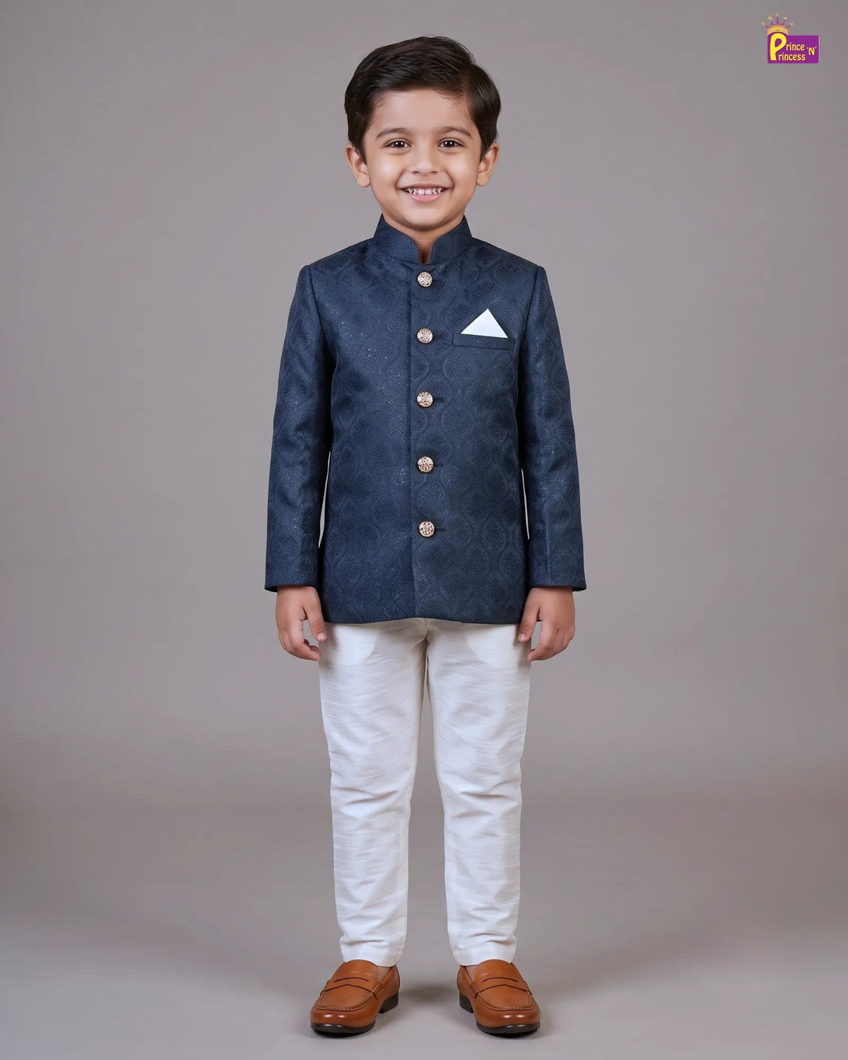 A young boy modeling a dark navy blue textured Indian sherpa jacket with gold buttons, paired with white trousers and brown leather shoes, standing against a neutral gray studio background.