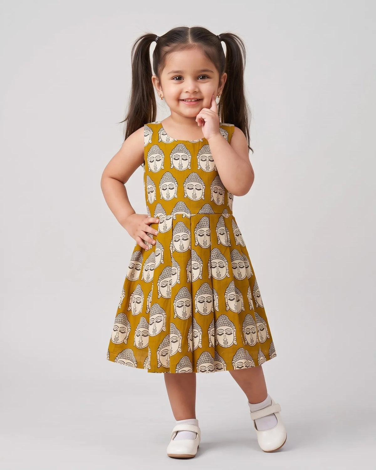 A young girl with dark hair in pigtails stands against a light gray studio background wearing a sleeveless mustard yellow dress with an all-over beige Buddha face print, white socks, and white shoes.