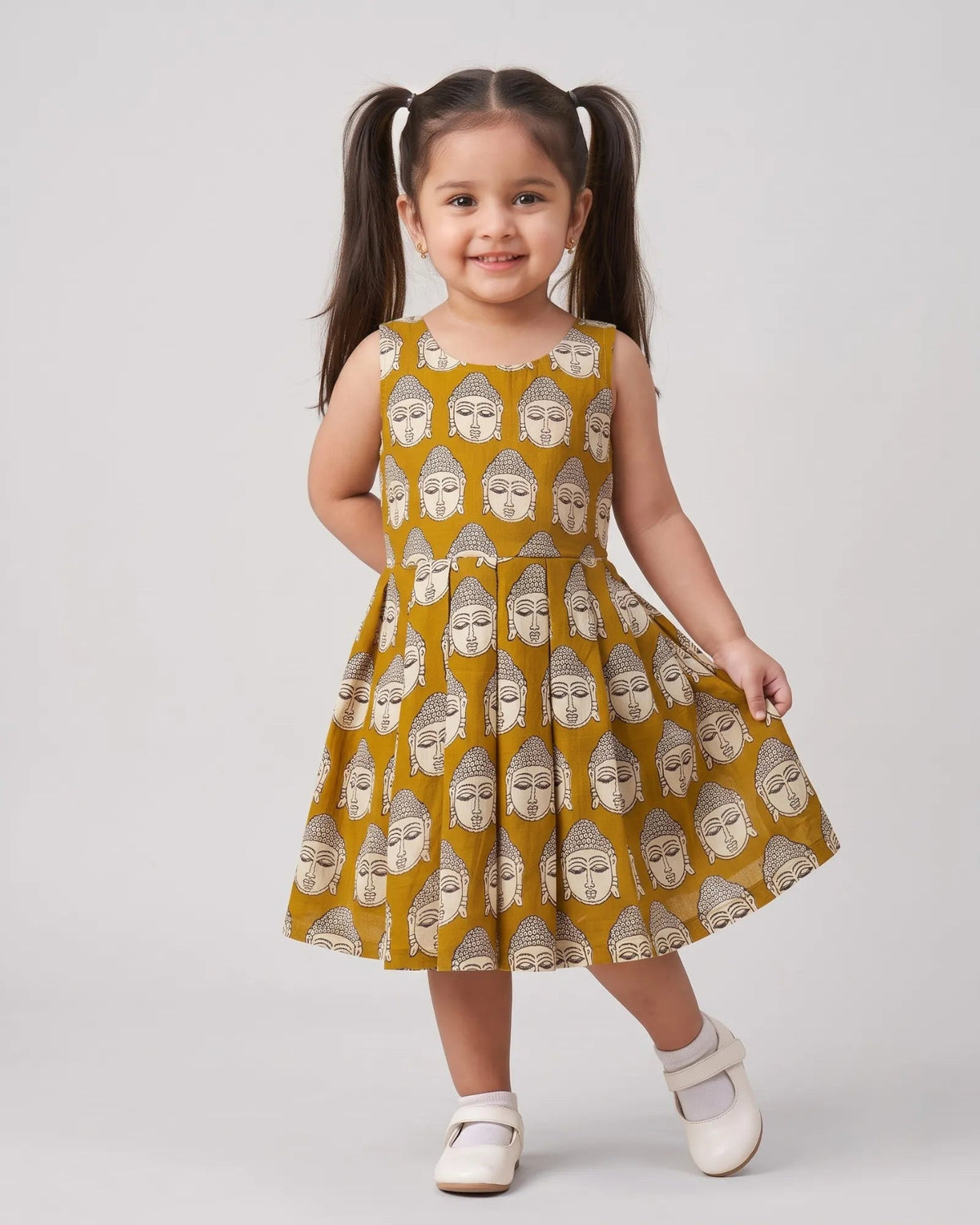 A young girl with dark brown pigtails stands in a sleeveless yellow dress with a repeating gray Buddha face pattern, paired with white shoes, against a light gray studio background.