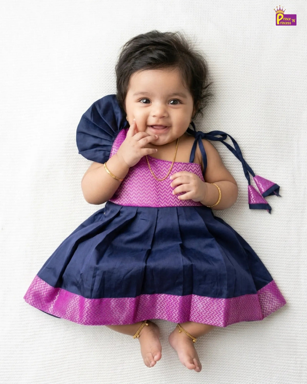A baby girl lying on a white textured blanket, wearing a navy blue and magenta traditional Indian dress with gold jewelry.